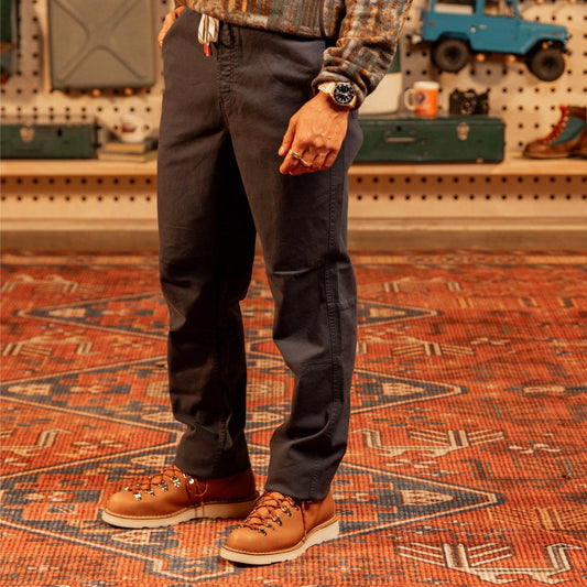 General image of person wearing dark pants and brown boots standing on a patterned rug with a workshop background.