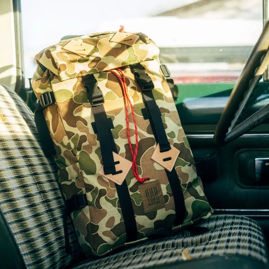Duck camouflage backpack with black straps and red cord detail, positioned on a car seat.