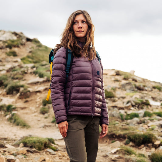 General image of woman in a purple jacket and green backpack hiking on a rocky mountain trail.
